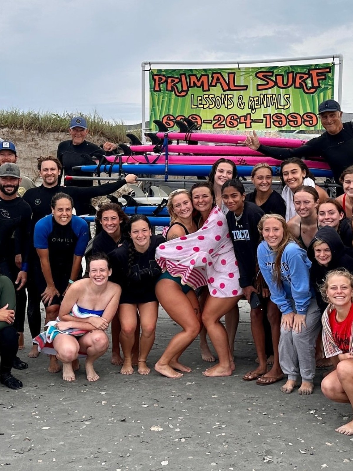 Group of people in beachwear posing on sand with surfboards and an SUV.