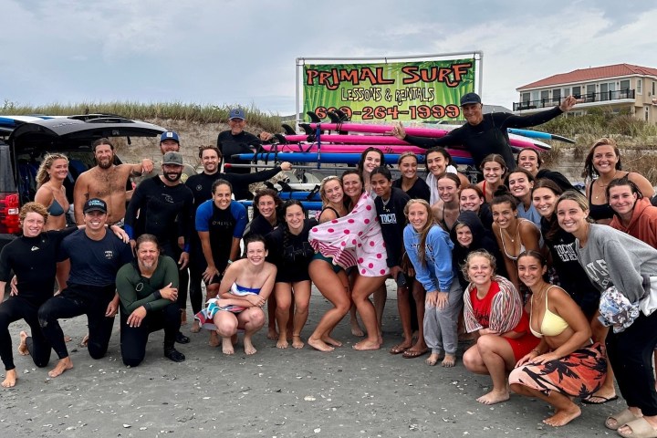 Group of people in beachwear posing on sand with surfboards and an SUV.