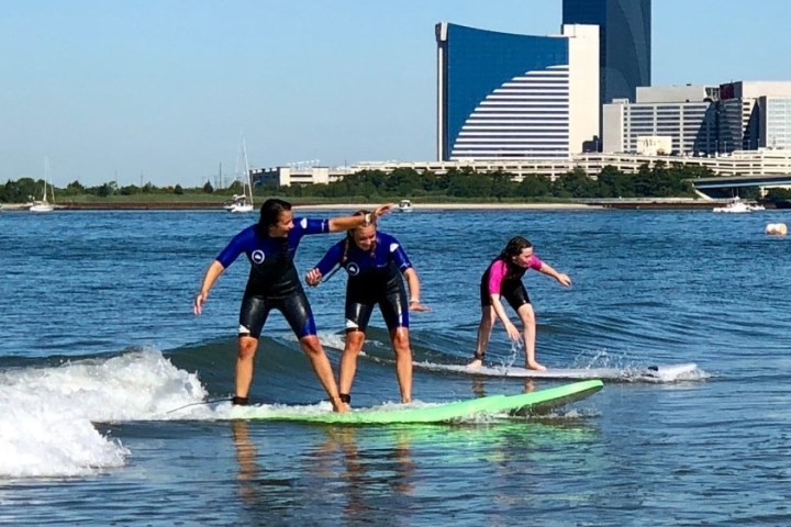 Three people surfing on a calm river with city skyline in the background.