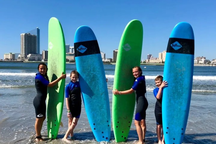 Four people in wetsuits holding surfboards on a beach with city skyline.