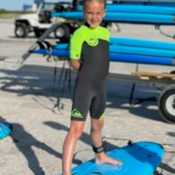 Child in wetsuit stands on blue surfboard at beach.