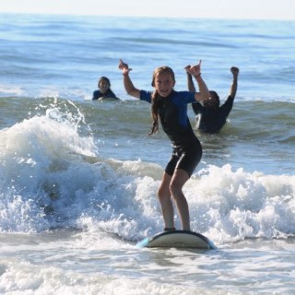 Young person in wetsuit surfing on waves, others in background.